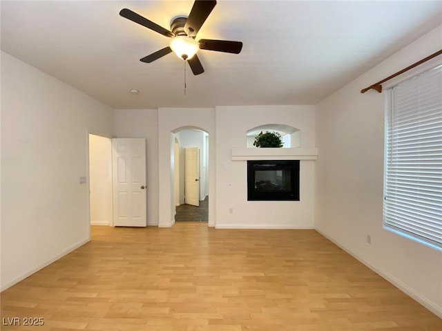 unfurnished living room featuring arched walkways, a ceiling fan, a glass covered fireplace, light wood-type flooring, and baseboards