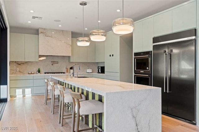 kitchen featuring built in fridge, backsplash, light wood-style floors, a kitchen island with sink, and a sink