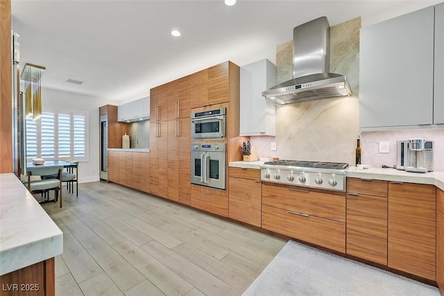 kitchen featuring brown cabinetry, light countertops, wall chimney range hood, and appliances with stainless steel finishes