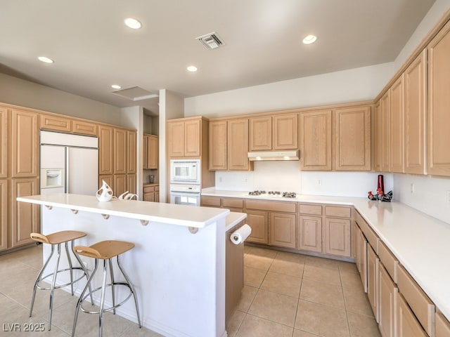 kitchen featuring light brown cabinetry, visible vents, under cabinet range hood, built in appliances, and a breakfast bar area