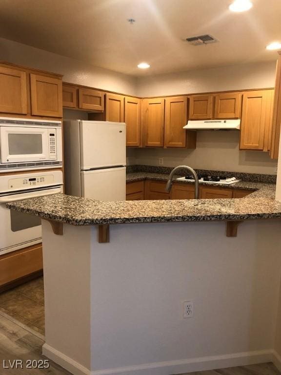 kitchen featuring white appliances, dark wood-style floors, dark stone countertops, under cabinet range hood, and a kitchen bar