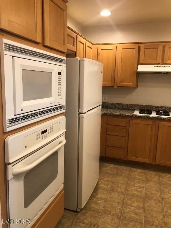 kitchen featuring brown cabinetry, dark countertops, white appliances, and under cabinet range hood