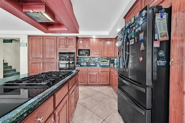 kitchen with brown cabinets, black appliances, a tray ceiling, tasteful backsplash, and light tile patterned floors