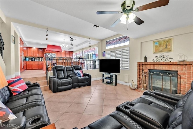 living area featuring baseboards, visible vents, light tile patterned flooring, a brick fireplace, and ceiling fan with notable chandelier