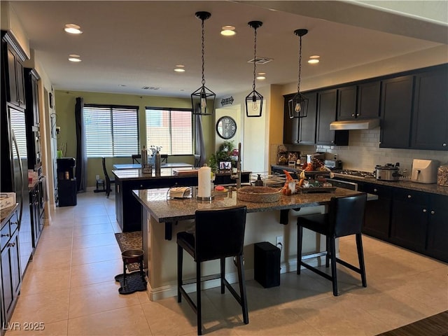 kitchen with light tile patterned floors, tasteful backsplash, a kitchen island with sink, dark stone countertops, and a kitchen breakfast bar