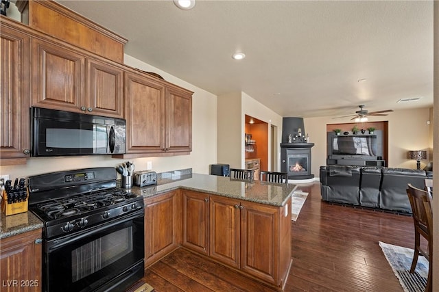 kitchen featuring dark wood-type flooring, dark stone countertops, a warm lit fireplace, a peninsula, and black appliances