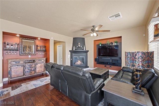 living area featuring ceiling fan, dark wood-style flooring, visible vents, baseboards, and a glass covered fireplace