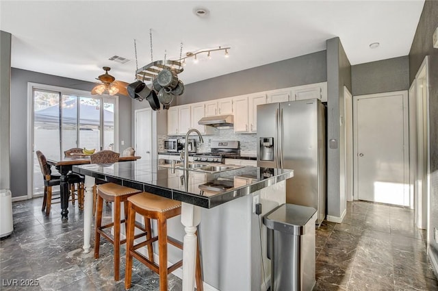 kitchen with under cabinet range hood, stainless steel appliances, visible vents, a kitchen breakfast bar, and tasteful backsplash