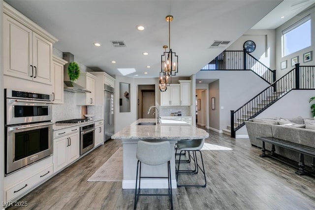 kitchen featuring visible vents, an island with sink, wall chimney exhaust hood, appliances with stainless steel finishes, and open floor plan