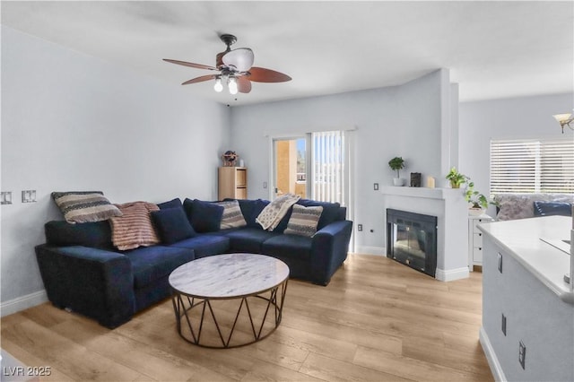 living room featuring a ceiling fan, a glass covered fireplace, light wood-style flooring, and baseboards