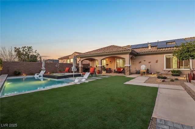 back of property at dusk with a patio, a fenced backyard, a yard, roof mounted solar panels, and stucco siding