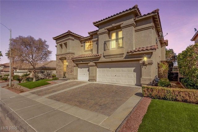 mediterranean / spanish house featuring an attached garage, a balcony, stucco siding, a tiled roof, and decorative driveway