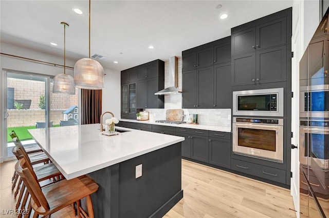 kitchen featuring a breakfast bar area, a sink, light countertops, appliances with stainless steel finishes, and wall chimney exhaust hood