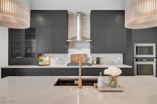 kitchen featuring glass insert cabinets, stainless steel appliances, light countertops, wall chimney range hood, and backsplash