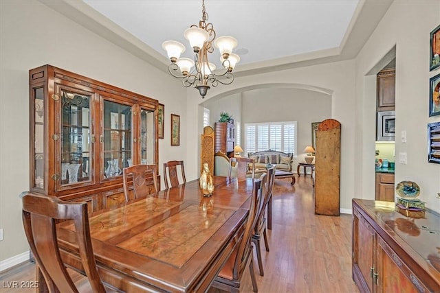 dining area with arched walkways, light wood-style flooring, a notable chandelier, baseboards, and a tray ceiling