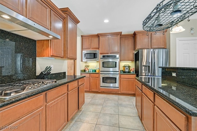 kitchen with light tile patterned floors, appliances with stainless steel finishes, brown cabinets, dark stone countertops, and under cabinet range hood