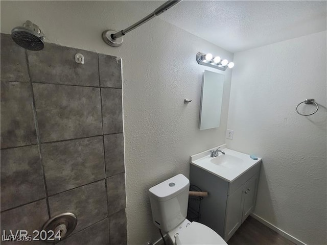 bathroom with toilet, a textured wall, tiled shower, and vanity