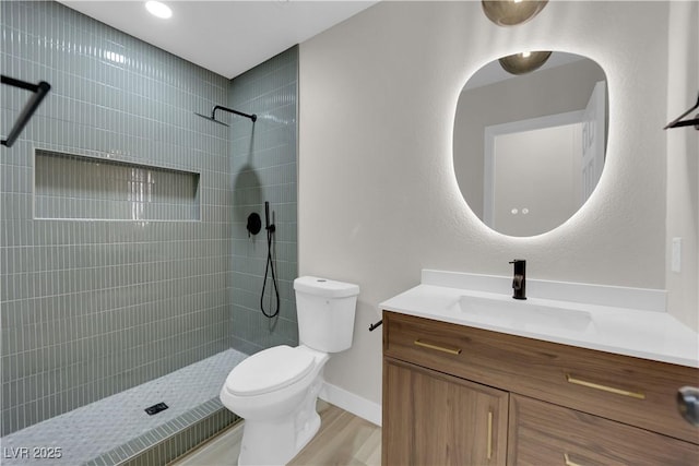 bathroom with toilet, wood finished floors, vanity, baseboards, and tiled shower