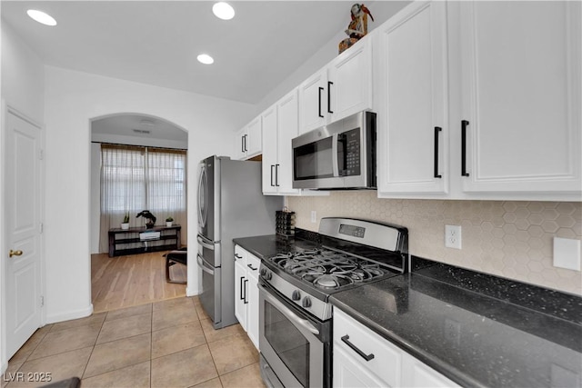 kitchen featuring arched walkways, light tile patterned flooring, stainless steel appliances, white cabinetry, and dark stone counters