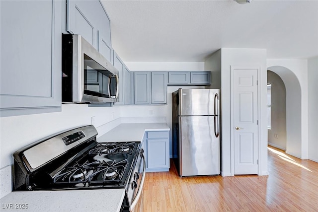 kitchen featuring arched walkways, light wood-style flooring, gray cabinets, stainless steel appliances, and light countertops