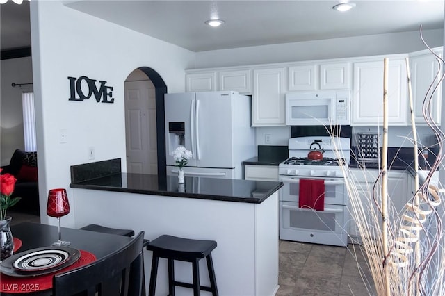 kitchen with a peninsula, white appliances, a breakfast bar, white cabinetry, and dark countertops