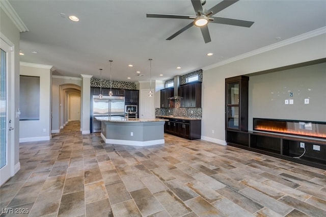 kitchen with backsplash, a glass covered fireplace, open floor plan, an island with sink, and wall chimney exhaust hood