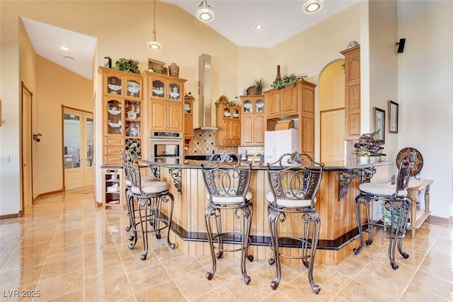 kitchen with a breakfast bar, a towering ceiling, freestanding refrigerator, stainless steel oven, and wall chimney range hood