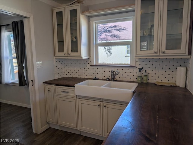 kitchen featuring a sink, baseboards, decorative backsplash, dark wood finished floors, and glass insert cabinets