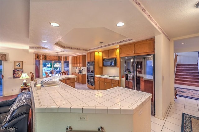 kitchen featuring black appliances, a sink, a tray ceiling, brown cabinetry, and tile counters