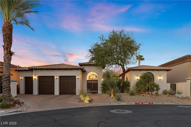 mediterranean / spanish house with an attached garage, a tile roof, decorative driveway, and stucco siding