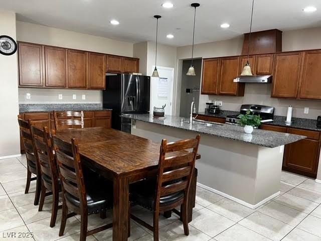 kitchen featuring stainless steel stove, brown cabinetry, an island with sink, under cabinet range hood, and black fridge