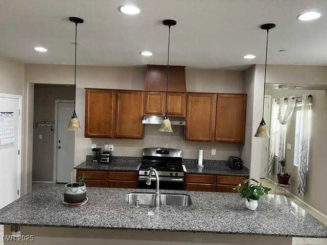 kitchen featuring stainless steel gas range oven, brown cabinets, dark stone countertops, under cabinet range hood, and a sink