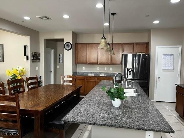 kitchen with light tile patterned flooring, a sink, dark stone countertops, an island with sink, and black fridge