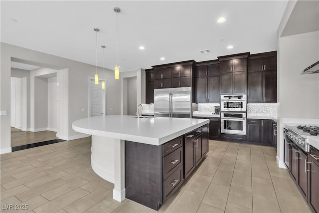 kitchen with light countertops, backsplash, appliances with stainless steel finishes, a kitchen island with sink, and dark brown cabinets