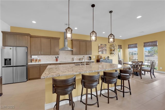 kitchen featuring stainless steel refrigerator with ice dispenser, decorative backsplash, a sink, light stone countertops, and wall chimney exhaust hood