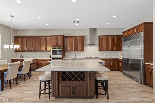 kitchen featuring wall chimney range hood, stainless steel appliances, an inviting chandelier, and a kitchen breakfast bar