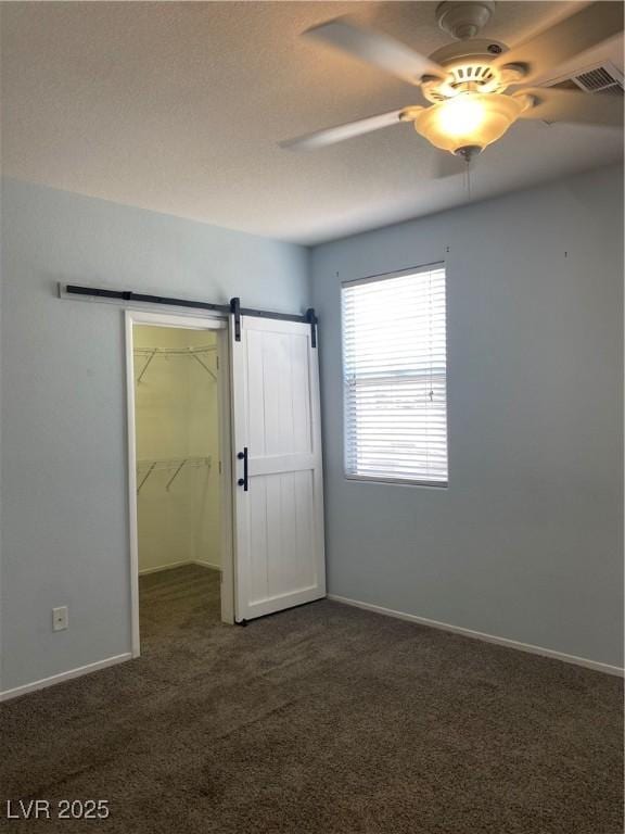 unfurnished bedroom with dark colored carpet, visible vents, a spacious closet, a barn door, and a ceiling fan