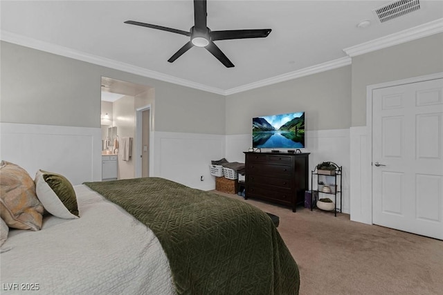 carpeted bedroom featuring ensuite bathroom, a wainscoted wall, a ceiling fan, visible vents, and crown molding