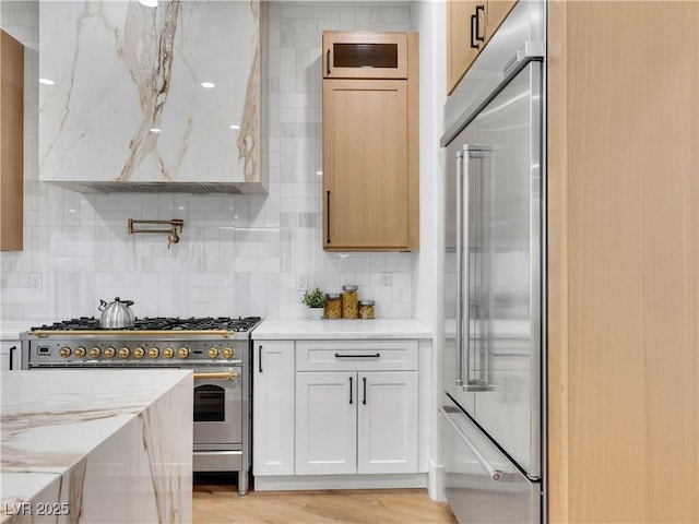 kitchen with light stone counters, exhaust hood, high quality appliances, backsplash, and light wood finished floors