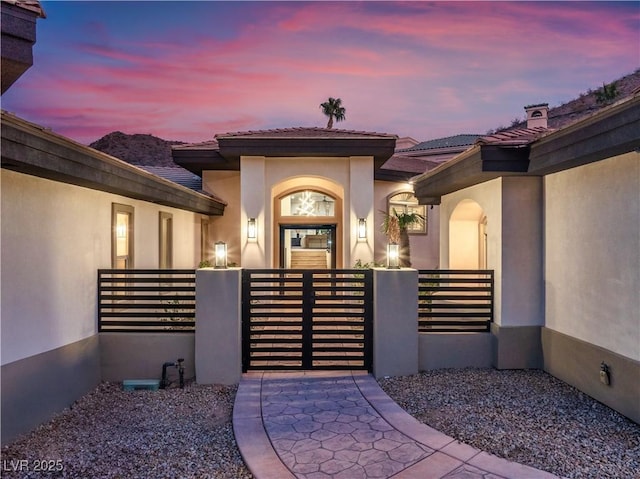 exterior entry at dusk with a tile roof, fence, a gate, and stucco siding