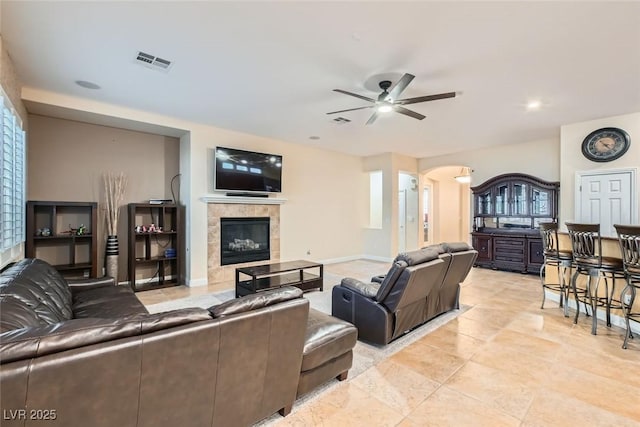 living area featuring arched walkways, visible vents, a tiled fireplace, a ceiling fan, and baseboards