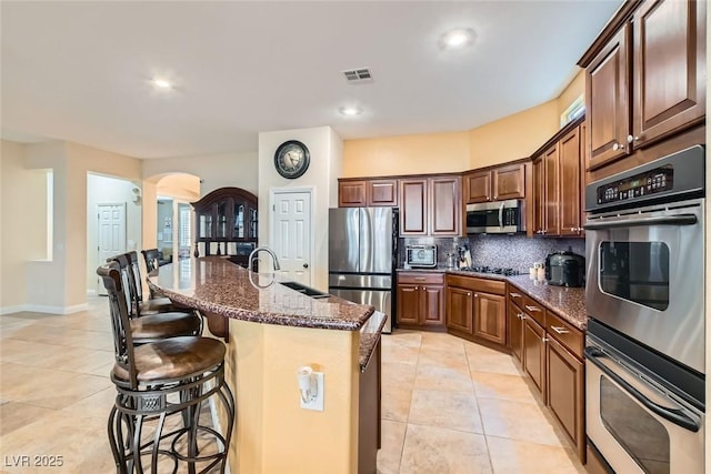 kitchen with arched walkways, stainless steel appliances, a kitchen breakfast bar, backsplash, and dark stone countertops