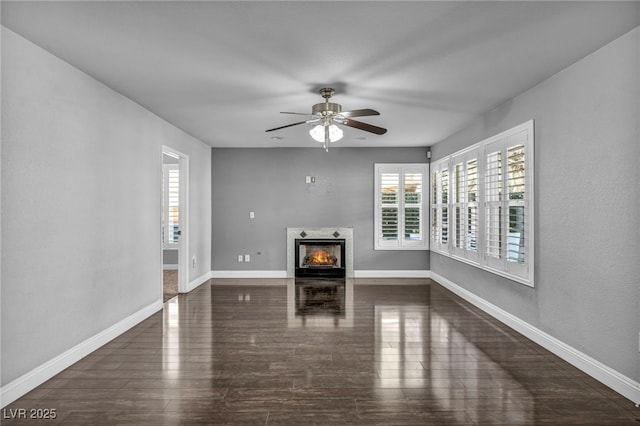 unfurnished living room featuring a warm lit fireplace, baseboards, dark wood-style floors, and a ceiling fan