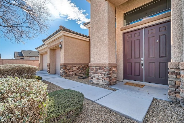 view of exterior entry with stone siding, a tiled roof, an attached garage, fence, and stucco siding