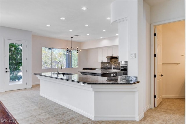 kitchen with a kitchen island with sink, a sink, white cabinetry, backsplash, and dark stone counters