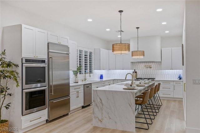 kitchen featuring light wood-style flooring, appliances with stainless steel finishes, hanging light fixtures, a kitchen island with sink, and backsplash