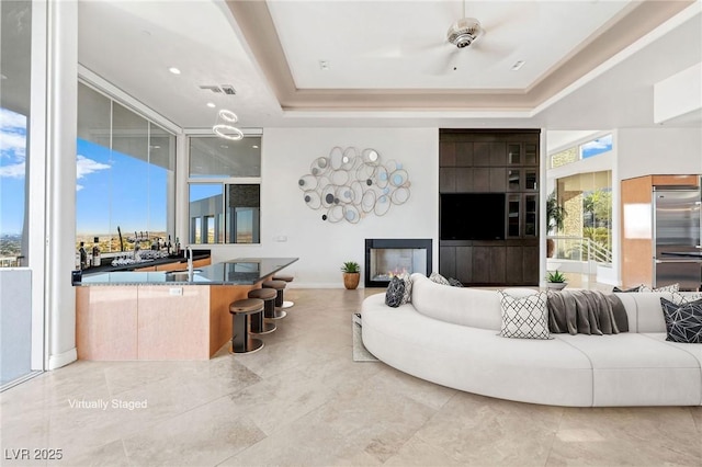 living area with baseboards, visible vents, a raised ceiling, a glass covered fireplace, and ceiling fan