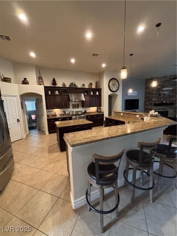 kitchen with arched walkways, a kitchen breakfast bar, dark brown cabinets, a large island with sink, and under cabinet range hood