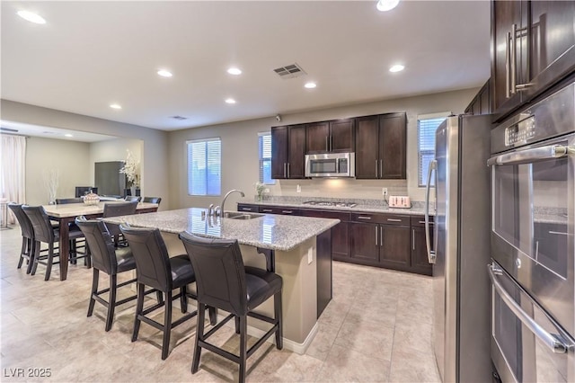 kitchen featuring a breakfast bar area, appliances with stainless steel finishes, a kitchen island with sink, a sink, and dark brown cabinets