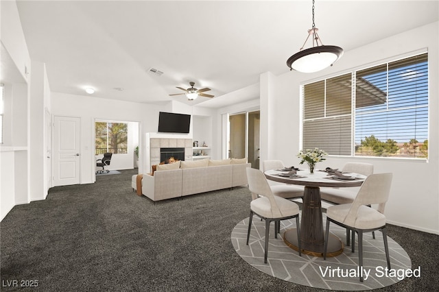 dining area featuring visible vents, dark carpet, a tiled fireplace, a ceiling fan, and baseboards